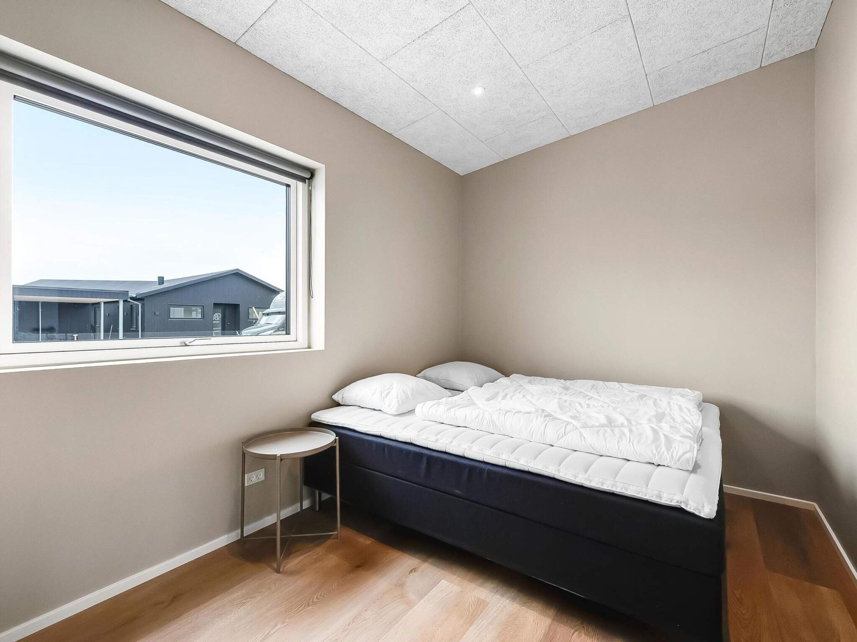 Un dormitorio con una cama, una mesita redonda pequeña, una ventana con vistas a edificios y un suelo de madera.