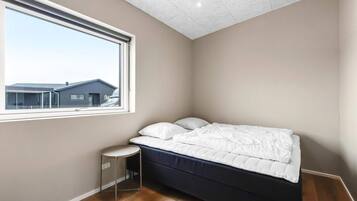 Un dormitorio con una cama, una mesita redonda pequeña, una ventana con vistas a edificios y un suelo de madera.