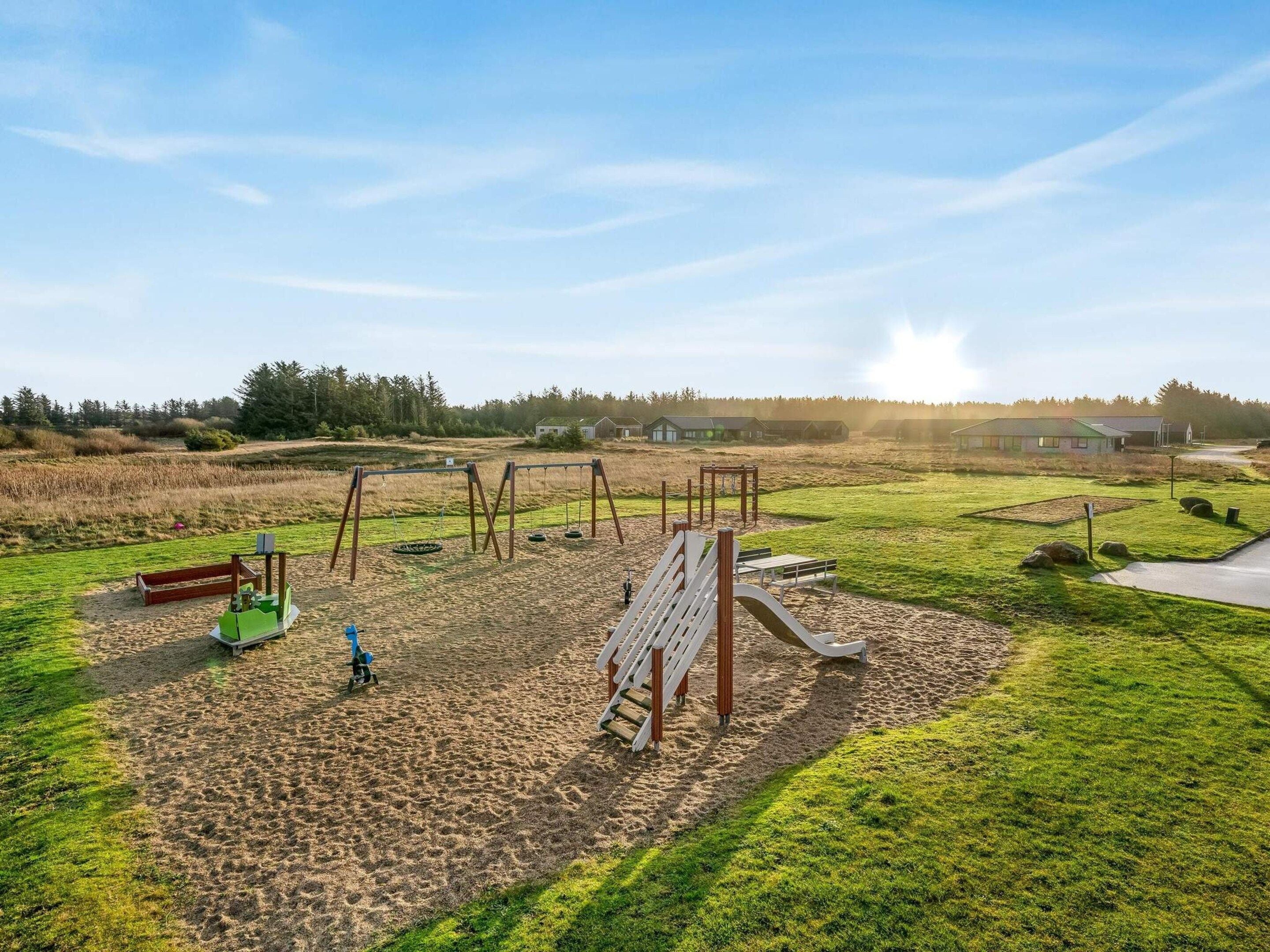 Un parque infantil con columpios y un tobogán, rodeado de zonas verdes y edificios al fondo.
