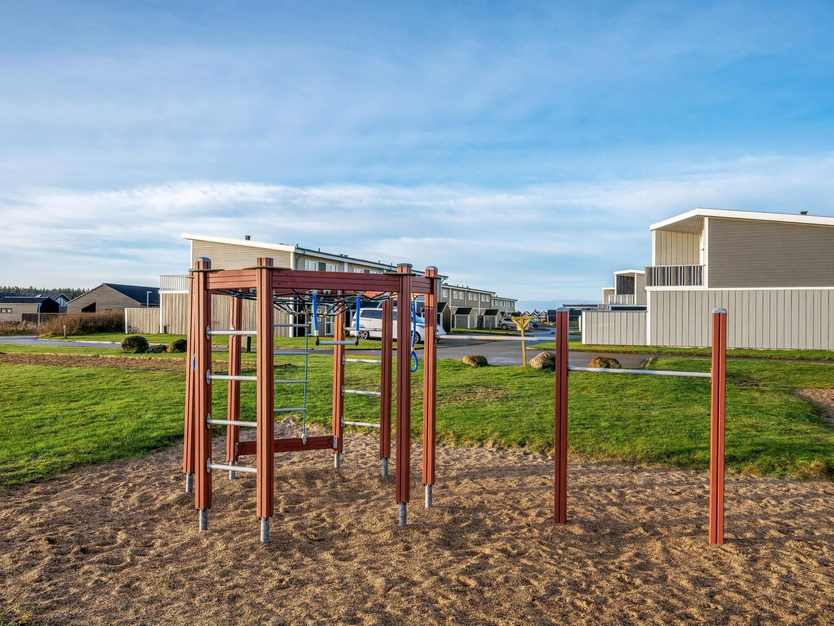 A playground with a climbing structure and swings, set against a backdrop of residential buildings and a clear sky.