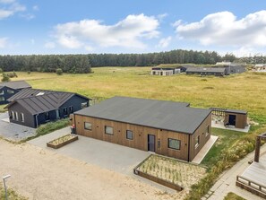 A modern building with a flat roof, surrounded by a grassy field and a forest in the background. - 4 Star Holiday Home in Lokken (Lokken)