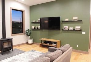 A living room with a flat-screen TV mounted on the wall, a sofa, a small wooden TV stand, a fireplace, and framed pictures on shelves.