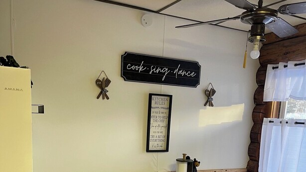 A kitchen with a white table and chairs, a black trash bin, a refrigerator, a ceiling fan, and a wall sign that reads "cook sing dance".