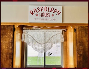 A window with a sign above it reading 'RASPBERRY HOUSE ESTABLISHED 2024' and a white lace curtain.