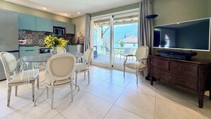 A dining area with a glass table and chairs, a TV on a wooden cabinet, and a kitchen with patterned tiles. - Grand T3 en Rez-de-jardin Tresserve (Tresserve)