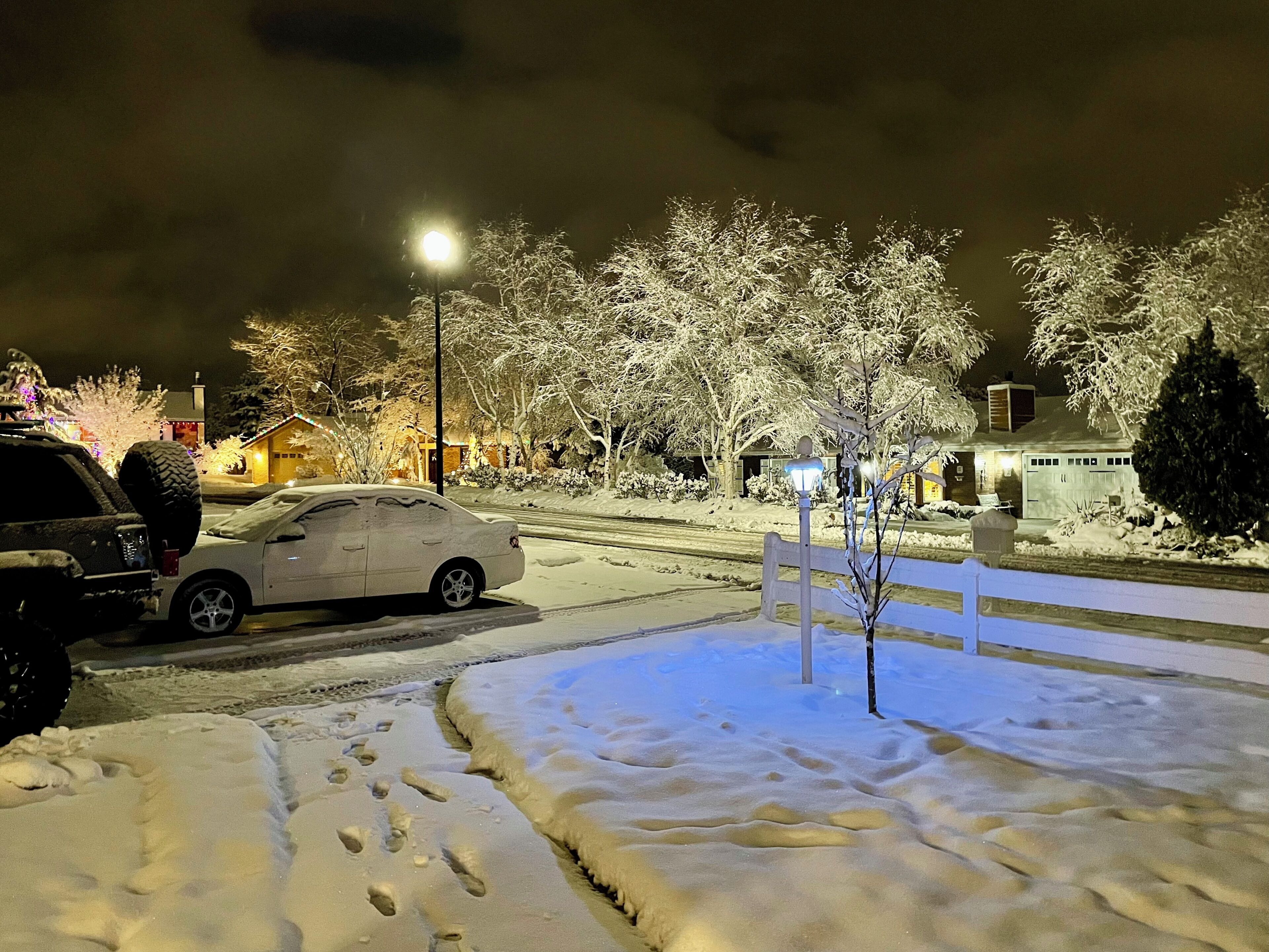 A snow-covered street at night with a parked car and a street lamp.