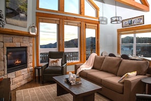 Ein gemütliches Wohnzimmer mit Kamin, Ledersessel, Sofa und großen Fenstern mit Blick ins Freie.