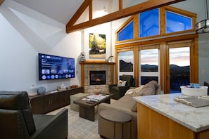 Ein Wohnzimmer mit Kamin, einem an der Wand befestigten Fernseher und Blick auf die Berge durch große Fenster.