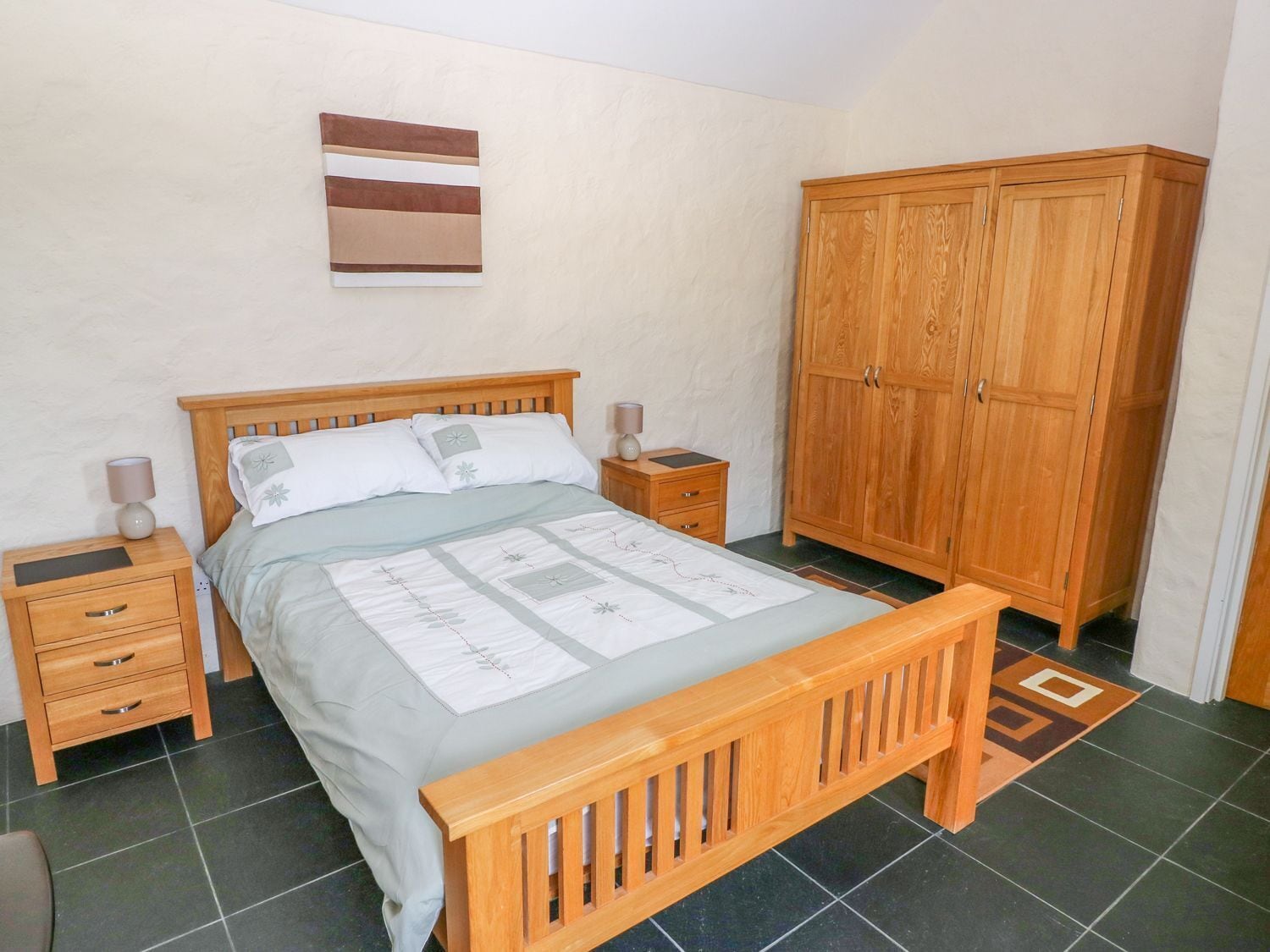 A bedroom with a wooden bed, two bedside tables with lamps, a wardrobe, and a wall-mounted artwork.