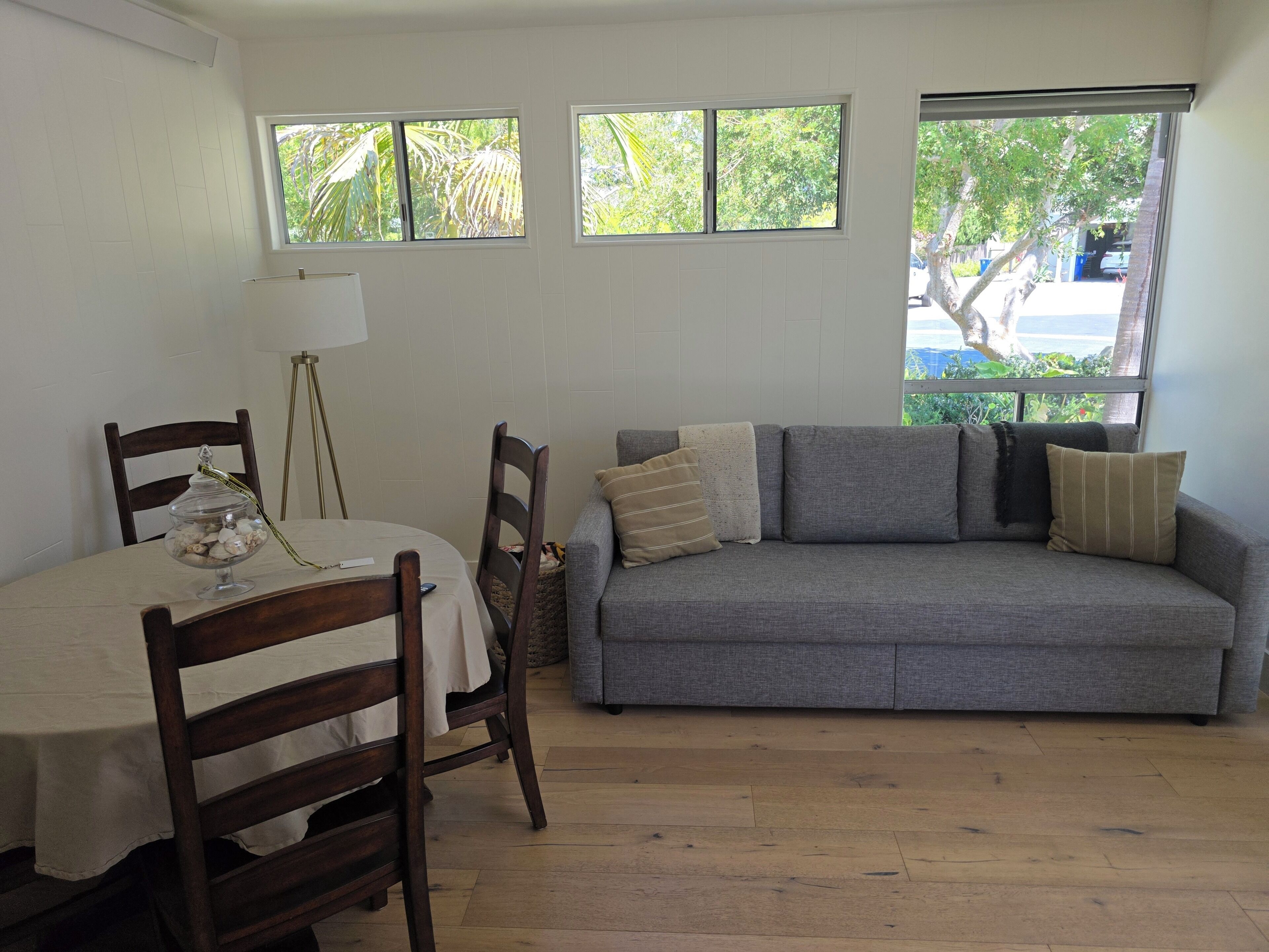 A room with a dining table, chairs, a sofa, and windows with a view of greenery.