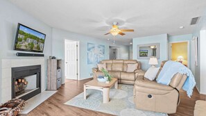 A living room with a fireplace, a ceiling fan, a television mounted on the wall, and a large window. - By The Lake Resort Access (Lake Ozark)