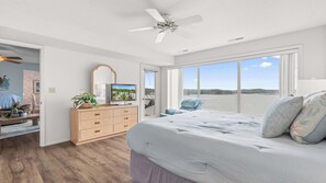 A bedroom with a large bed, a TV, a dresser, and a view of the water. - By The Lake Resort Access (Lake Ozark)