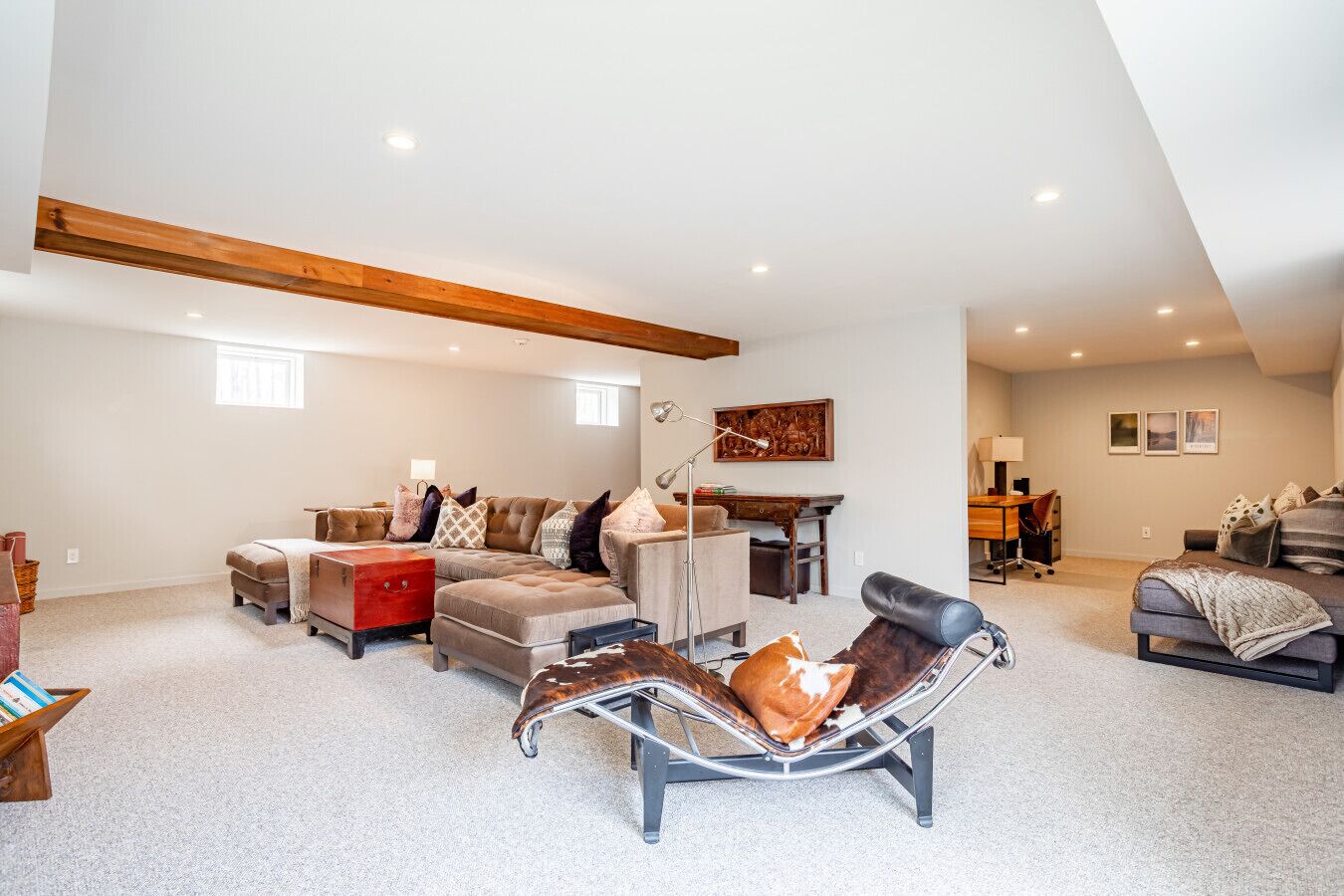 Un loft espacioso con techo alto, vigas de madera a la vista, un gran ventanal y una zona de estar moderna con sofá, reposapiés y una silla metálica original.