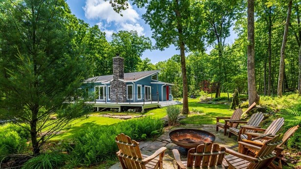 Ein Haus mit einem Steinkamin, umgeben von einem üppigen Garten und einer Sitzgelegenheit mit Adirondack-Stühlen.