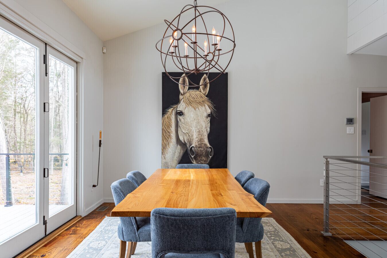 Een eetruimte met een houten tafel, blauwe stoelen, een kunstwerk met paardenthema en een moderne kroonluchter.