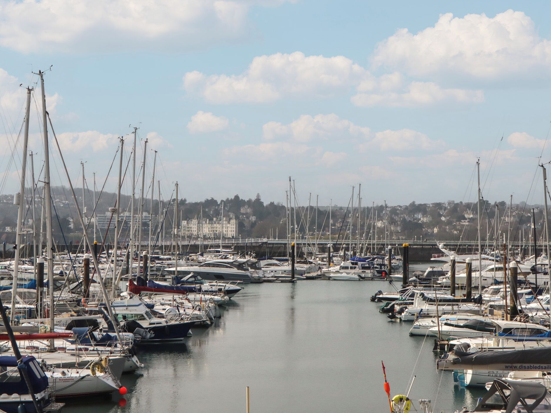 A marina with numerous boats docked, a calm water body, and a distant building complex.