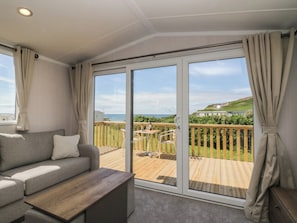 Una habitación con sofá, una mesa de madera y un ventanal corredero que da a una terraza exterior con vistas al mar y las colinas.