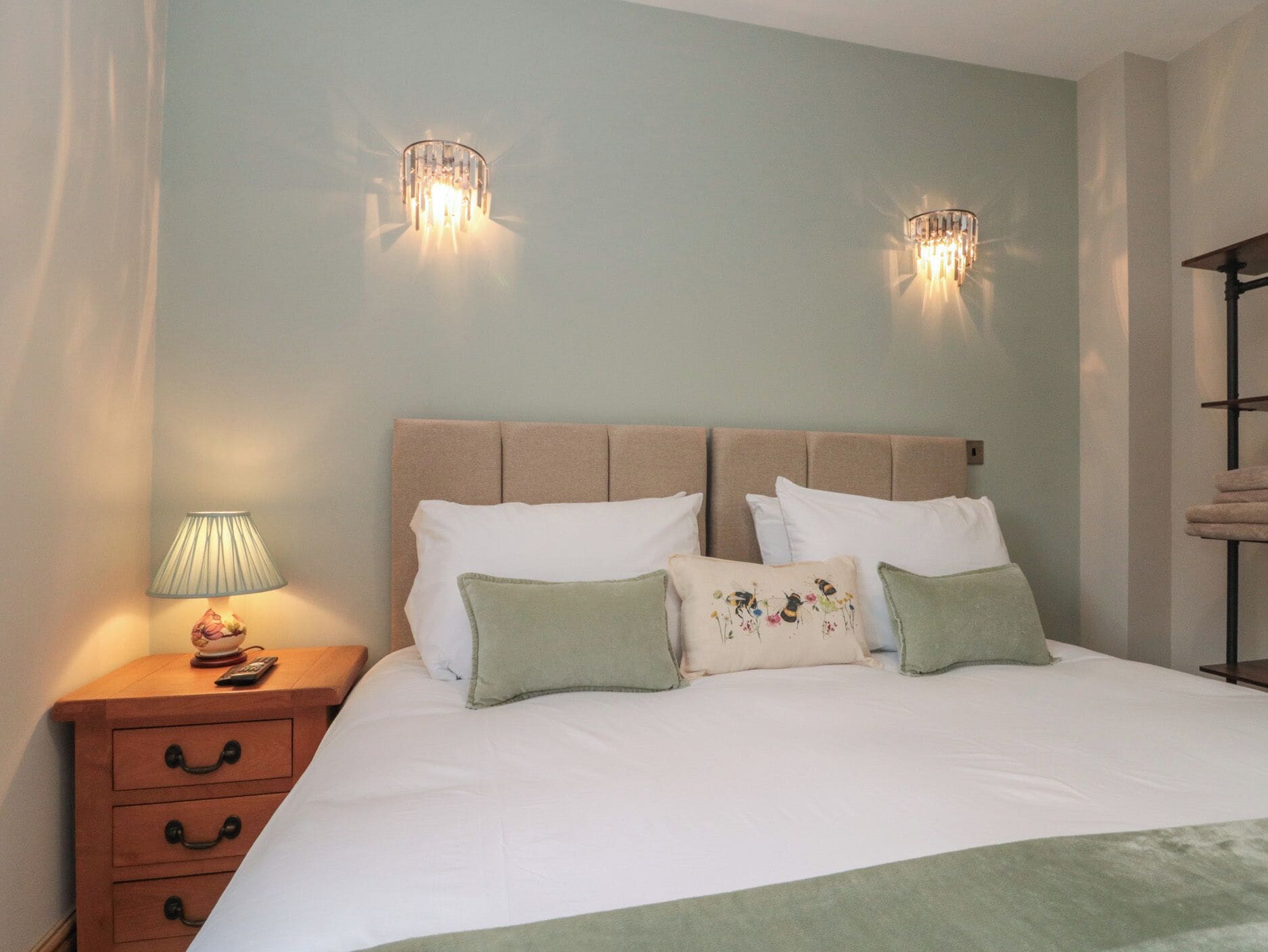 A neatly made bed with white linens and multiple pillows, a bedside table with a lamp, and two wall-mounted light fixtures.