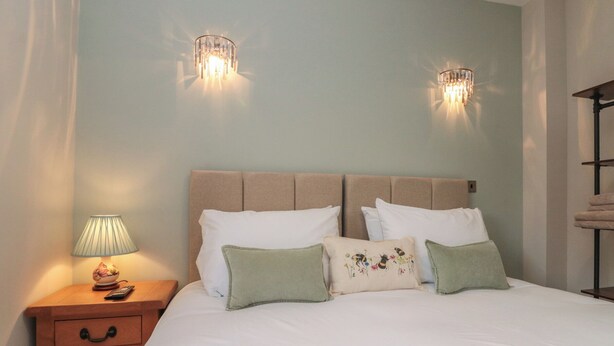 A neatly made bed with white linens and multiple pillows, a bedside table with a lamp, and two wall-mounted light fixtures. - Bumble Bee Lodge (Filey)