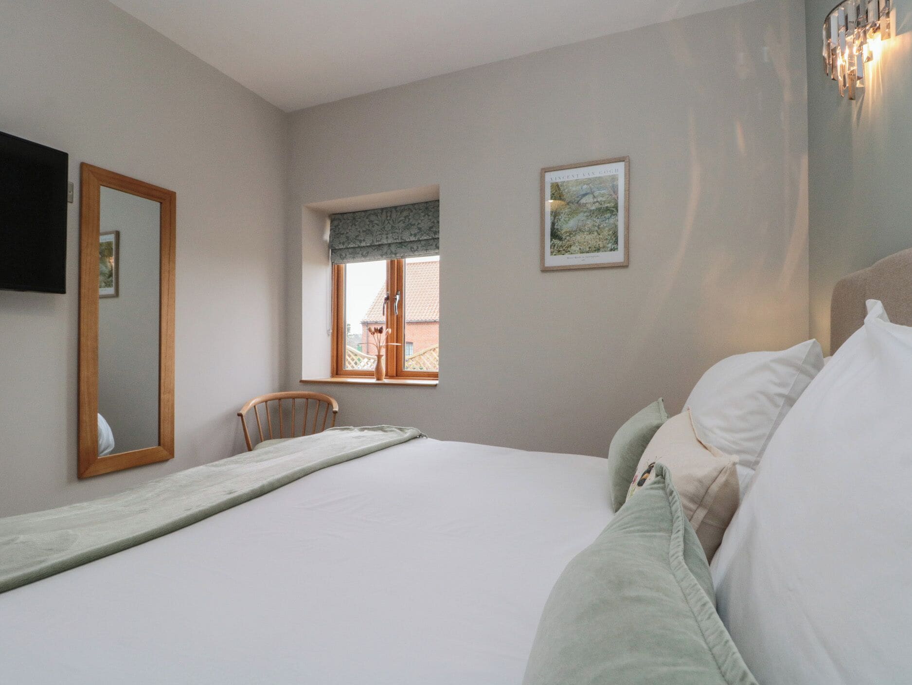 A bedroom with a bed, a window, a chair, a mirror, and a wall-mounted light fixture.