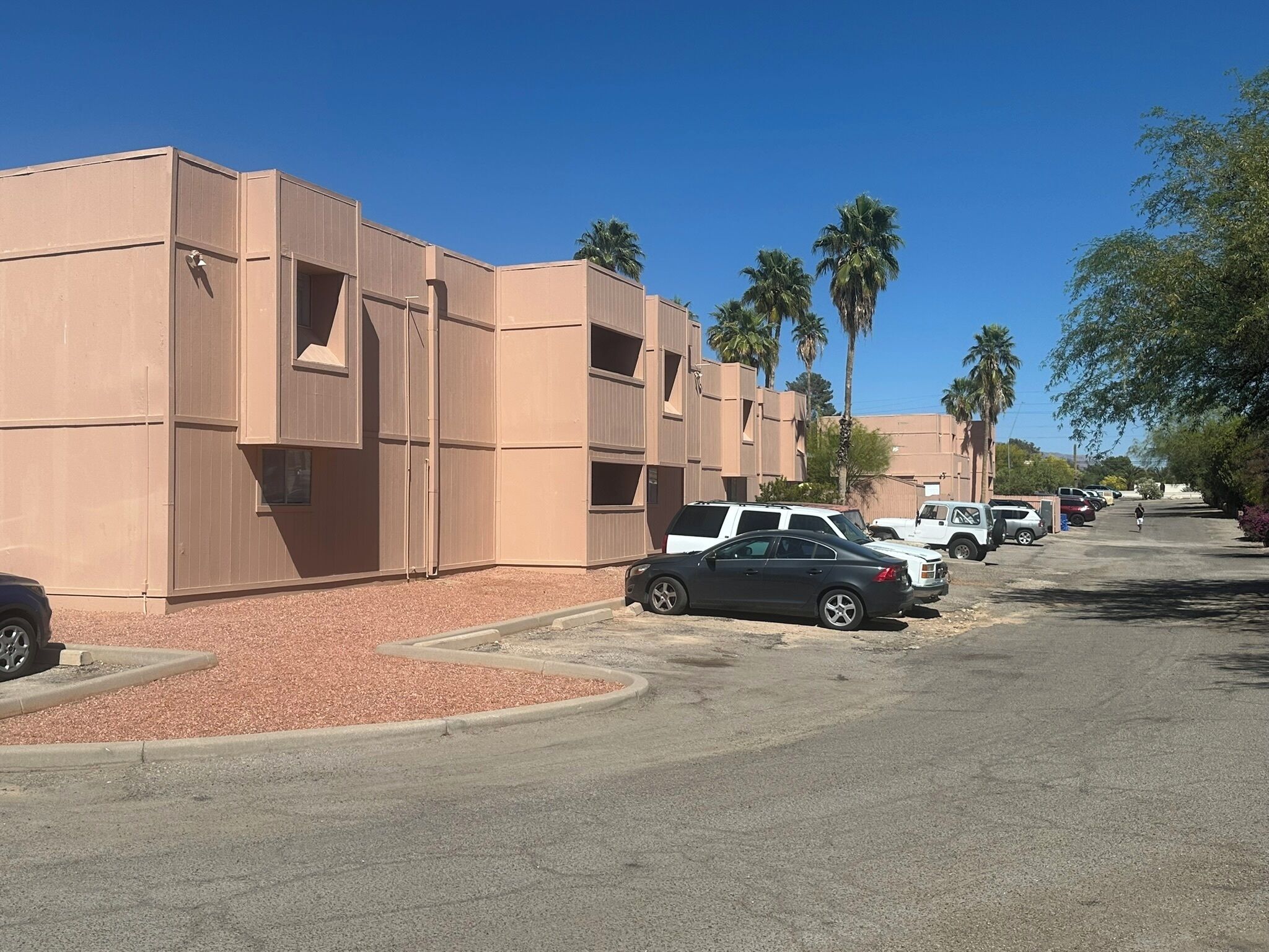A row of beige buildings with a parking lot in front, palm trees, and a clear blue sky.