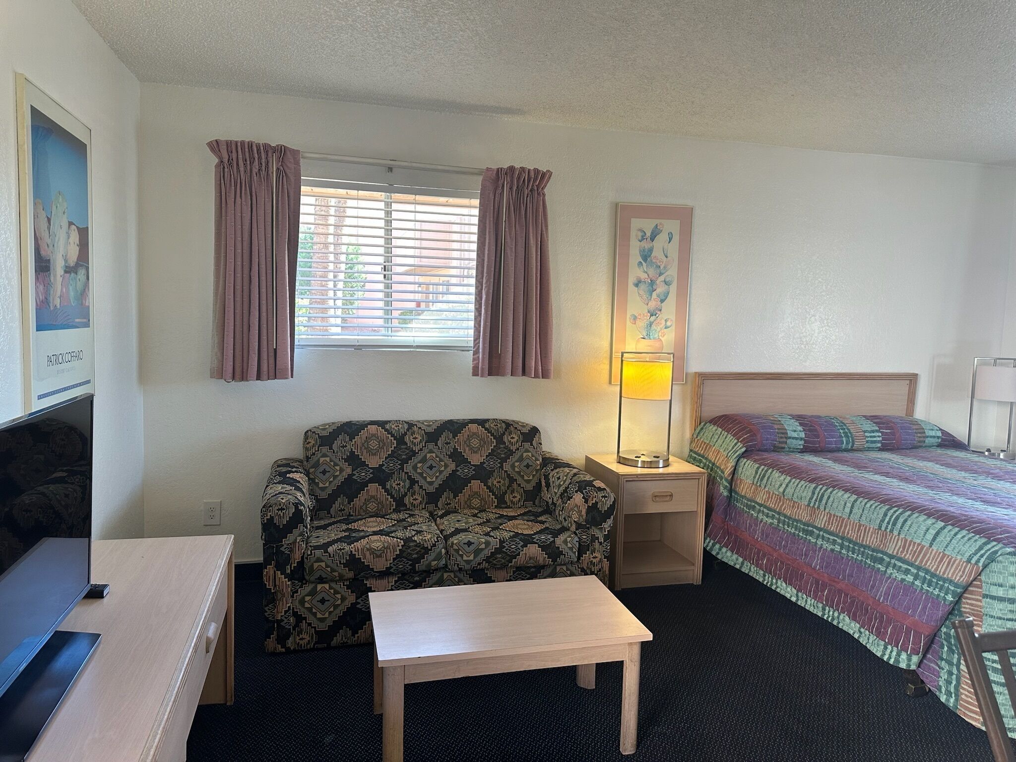 Habitación de hotel con sofá, una mesita, una mesita de noche con lámpara, cama con manta colorida y ventana con cortinas.