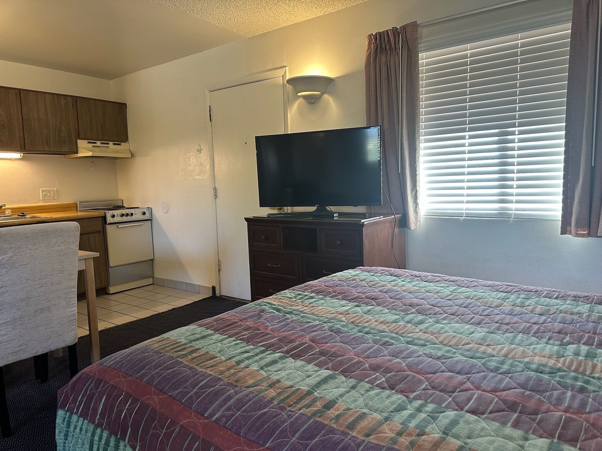 Une chambre d’hôtel compacte comprenant un coin cuisine, un lit avec une couverture aux couleurs vives, une télévision et une fenêtre avec des stores.