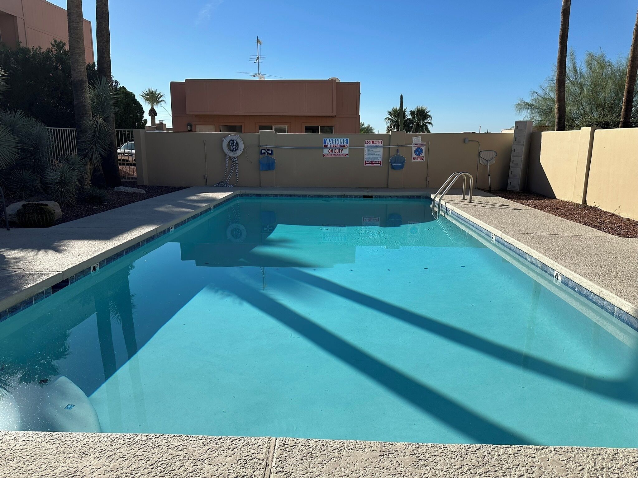 Une piscine aux eaux claires, avec un poste de surveillance pour les sauveteurs, un panneau indiquant les règles de la piscine et un autre panneau indiquant l’absence de sauveteur.