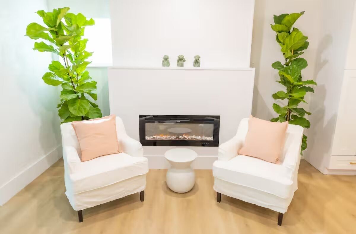 A room with two white armchairs, a small white table, and a fireplace with three small figurines on the mantel.