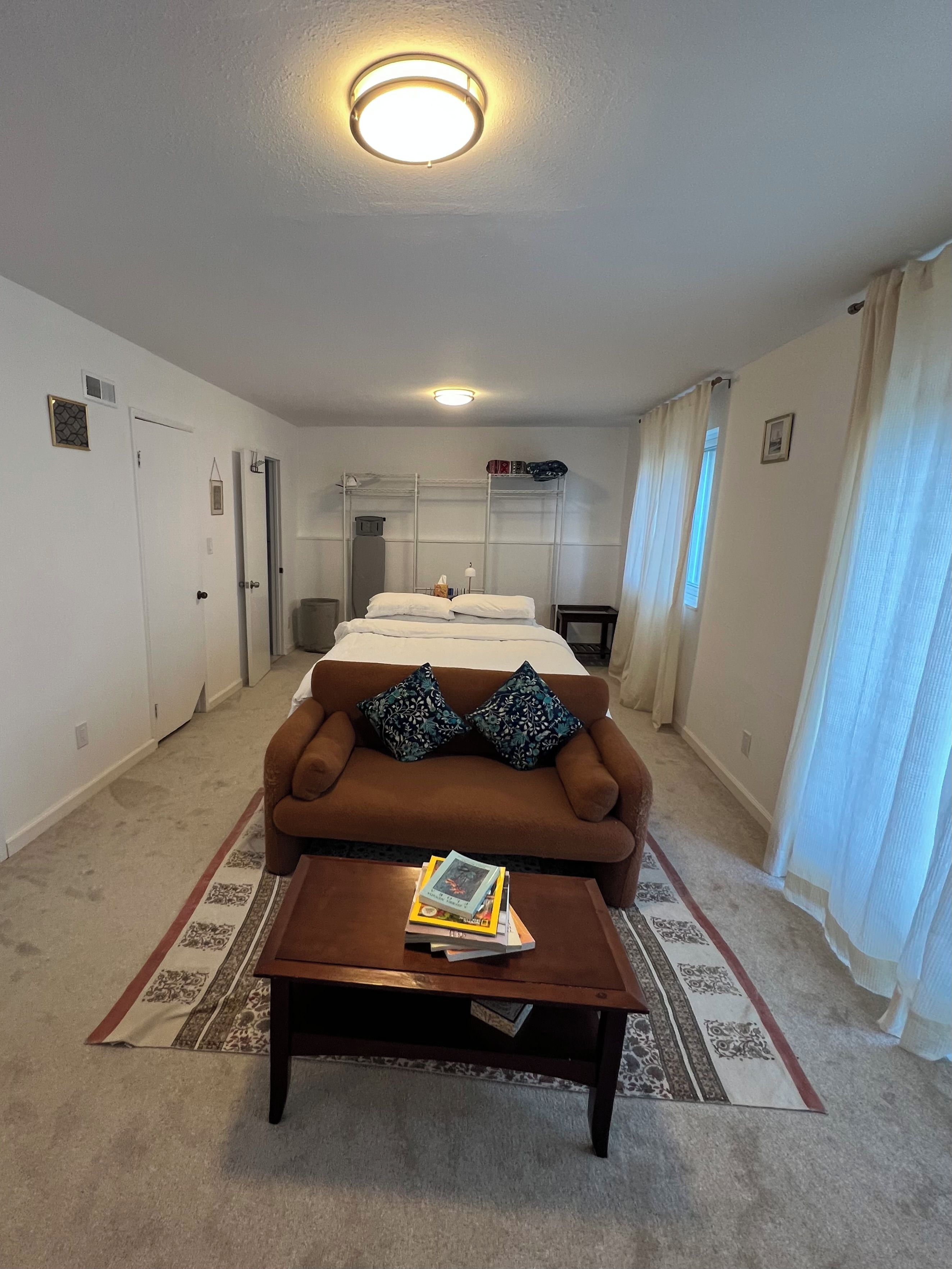 A hotel room with a brown sofa, a coffee table with books, a bed with white linens, and a ceiling light.