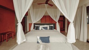 Una cama con dosel adornada con cortinas blancas, un ventilador de techo y un banco de madera.