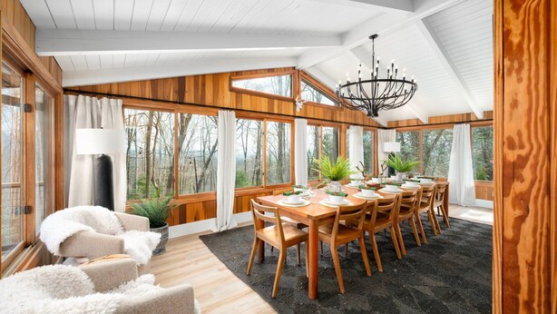 A dining area with a wooden table set for six, surrounded by chairs, with a chandelier above and large windows offering a view of trees. - Biltmore Ridgetop Escape | Hot Tub + Sauna (Asheville)