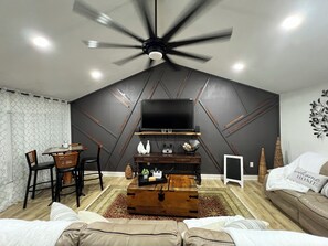 A modern living room with a ceiling fan, a TV, a wooden coffee table, a sofa, and a dining area with chairs. - Oak Canopy Cottage, Near Downtown, Lackland, & More (San Antonio)