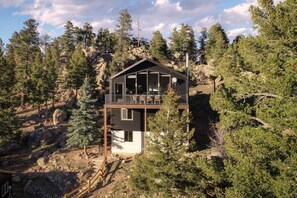 A two-story cabin with a balcony, surrounded by dense forest and rocky terrain. - Red Rocks Gather - Hot tub, sauna, cinema & views (Morrison)