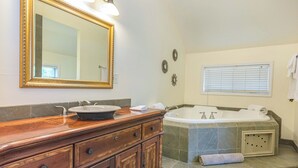 A bathroom with a wooden vanity, a large mirror, a bathtub, and a window with blinds. - Riverfront Suites • Fireplace • Whirlpool for Two • Walk to Downtown (Wisconsin Dells)