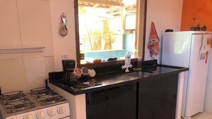 A compact kitchen with a gas stove, a coffee maker, a refrigerator, and a window with a view of greenery outside. - House 150 meters from the beach. (Paraty)