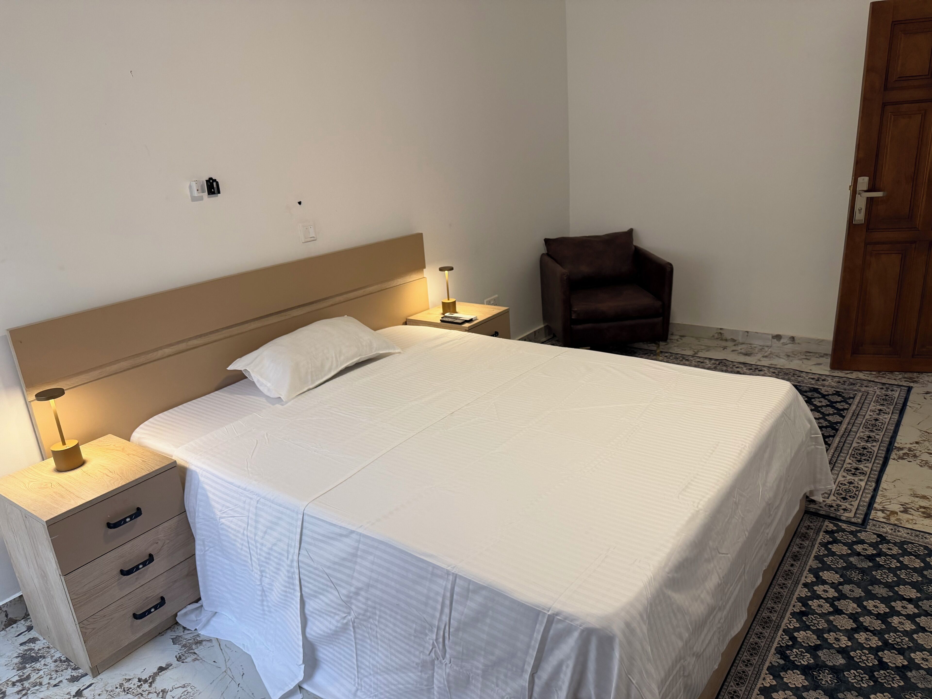 Una cama bien hecha con cabecera de madera, una mesita de noche con lámpara, una silla y un tapete estampado.