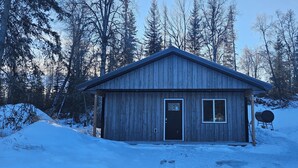 Una pequeña cabaña de madera con puerta y ventanas, rodeada de nieve y árboles.