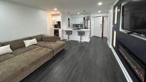 A modern living room with a sofa, a kitchen area with a bar, and a large TV. - Cozy 2-bedroom condo in quaint Virginia Beach with AC and gym (Virginia Beach)