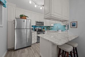 A modern kitchen with white cabinetry, stainless steel appliances, a marble countertop, and a bar stool. - Shipwatch 1105 - 2BR- 2BA - Oceanfront - Pool (North Topsail Beach)