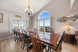 A dining area with a large table, chairs, a chandelier, and a view of the ocean through a large window. - 'A Sunny Delight - 6BR - Oceanfront - Sleeps 15 (North Topsail Beach)