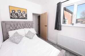Een slaapkamer met een geborduurde hoofdbord, een bed met witte beddengoed en gekleurde kussens, een raam met uitzicht op gebouwen, en een deur die leidt naar een andere kamer.