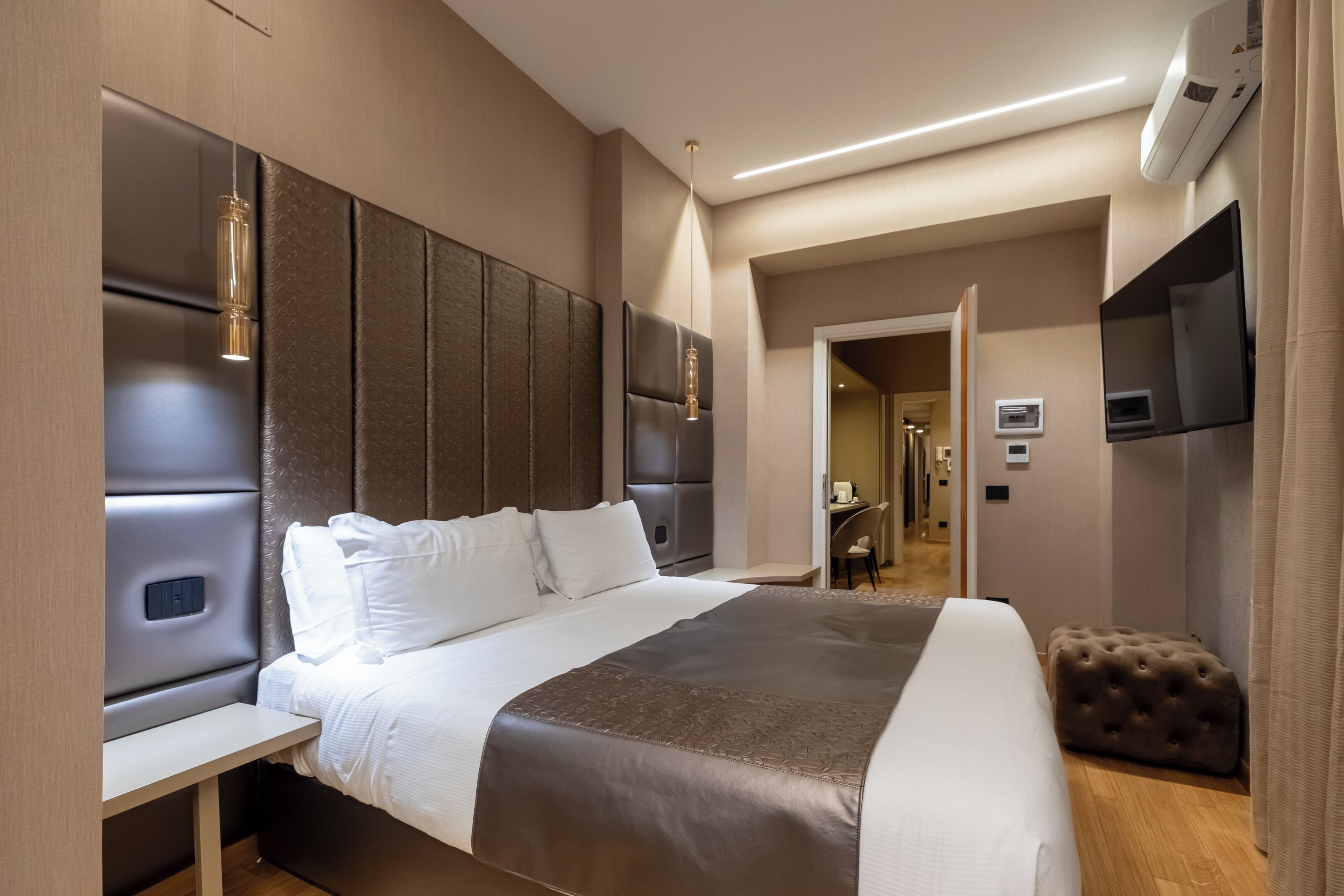 Une chambre d’hôtel moderne, dotée d’un grand lit, d’une table de chevet, d’un téléviseur fixé au mur et d’une porte donnant sur un couloir.