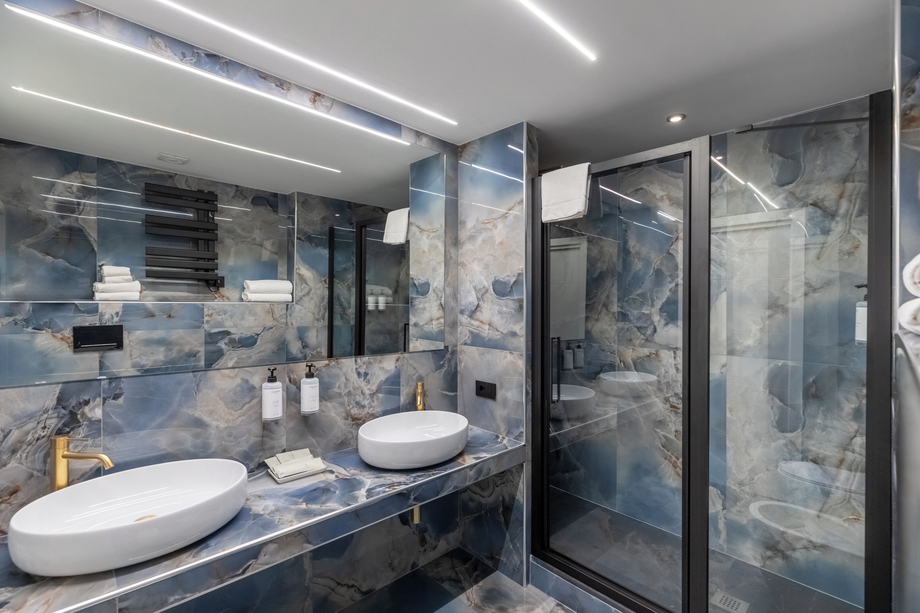 Une salle de bain moderne avec des murs en marbre, deux lavabos et une douche à l’italienne.