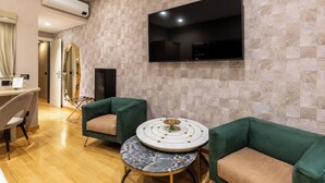 A modern living room with a flat-screen TV, two green armchairs, a round coffee table, and a marble accent table. - HELMIRAGE Frattina Suite (Rome)