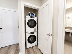 A laundry room with two stacked front-loading washing machines, a white door, and a visible bathroom with a sink and mirror. - Modern Loft Near Yale |Wooster Sq| 5 Star Location (New Haven)