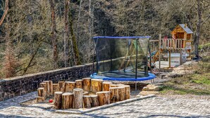 Un trampolín con red de seguridad, rodeado de troncos de madera, en un área boscosa con una casita de juegos de madera al fondo.