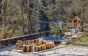 A trampoline with a safety net, surrounded by wooden stumps, in a wooded area with a wooden playhouse in the background. - Cozy apartment in Irrhausen with WiFi (Irrhausen)