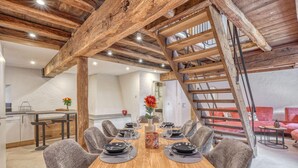 Un comedor con una mesa de madera dispuesta para seis personas, rodeada de sillas tapizadas de gris. Se observa una escalera de madera con barandilla de metal a la derecha.