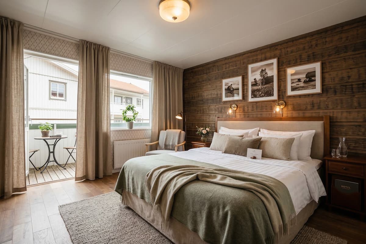 Une chambre d’hôtel avec un grand lit, une tête de lit en bois, une petite table avec des chaises, un balcon avec des rideaux et des tableaux encadrés au mur.